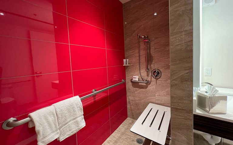 A bathroom with a red tiled wall, a shower with a grab bar and soap shelf, and a towel hanging on the wall.