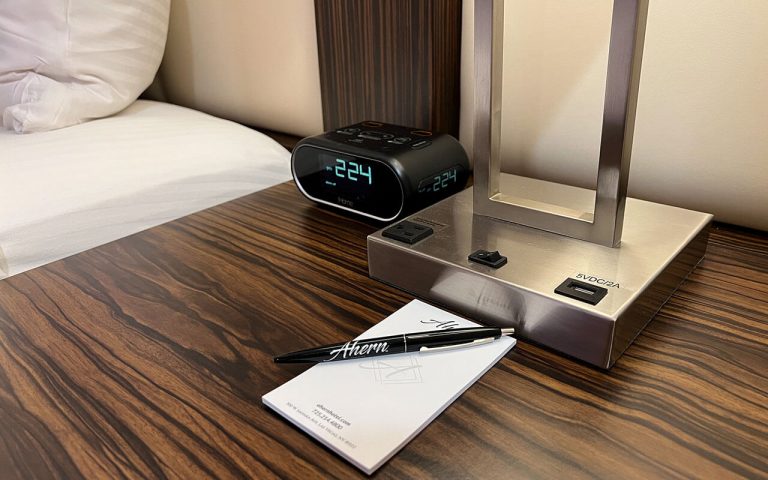 A bedside table with a digital alarm clock, notepad, and pen.
