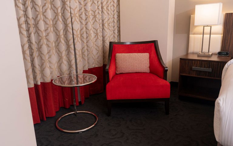 A red accent chair with a beige pillow sits in a hotel room next to a glass side table and a wooden nightstand with a lamp.