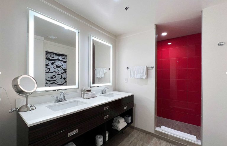 A modern bathroom with a double vanity, two large mirrors, and a red accent wall in the shower area.