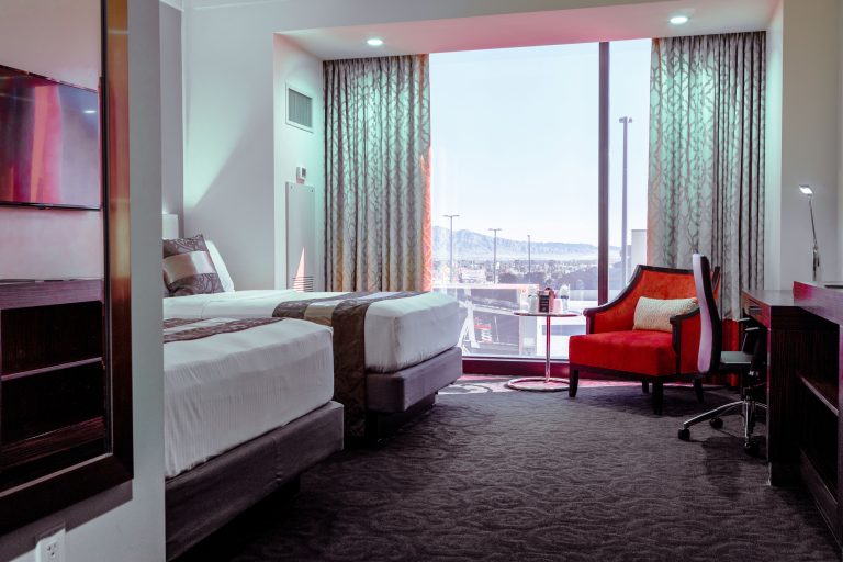 A hotel room with two beds, a desk, an armchair, and a large window.