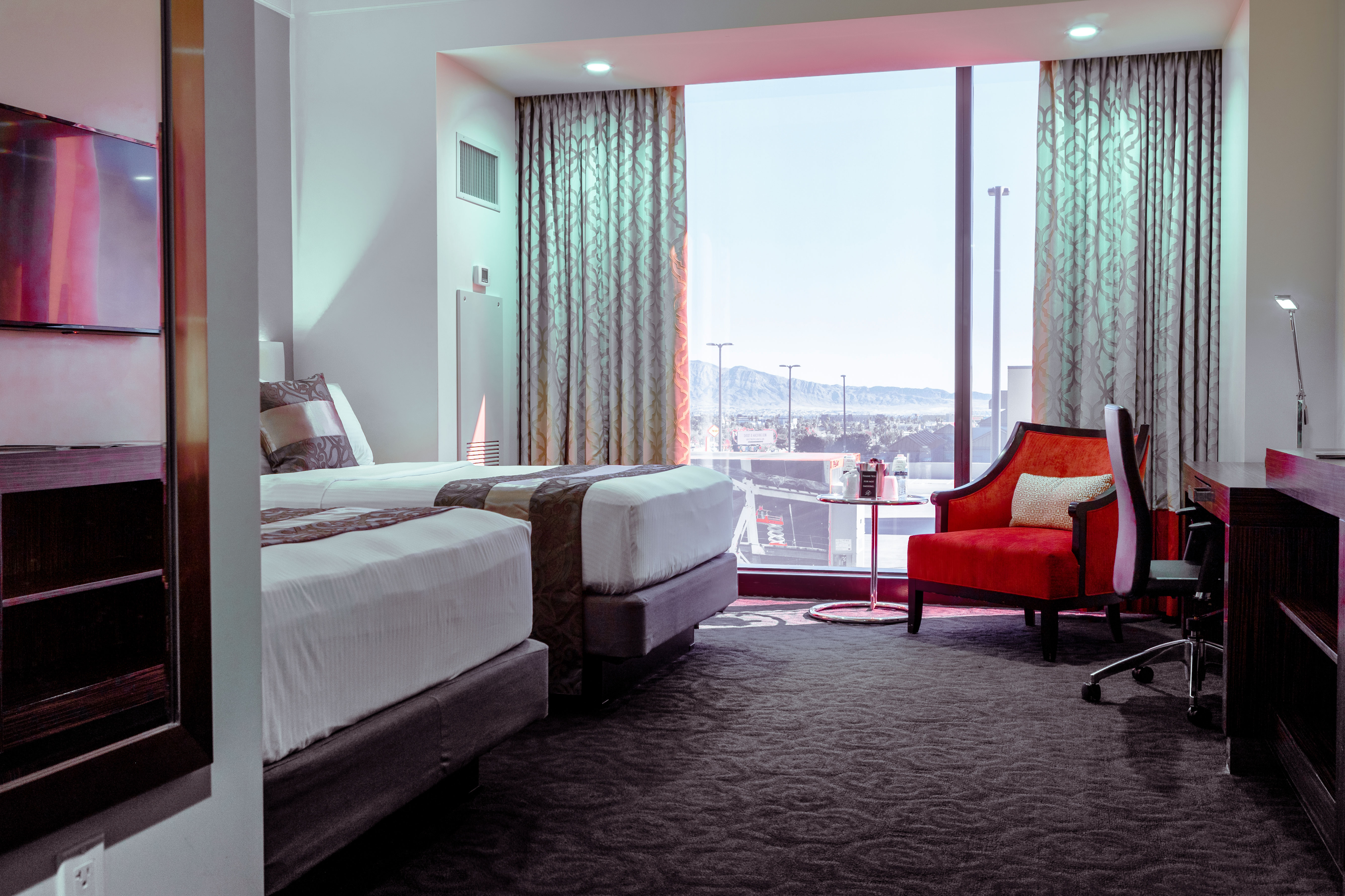 A hotel room with two beds, a desk, an armchair, and a large window.