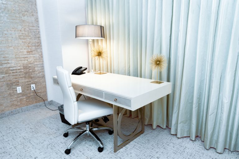 A white desk with a chair, lamp, and curtains in the background.